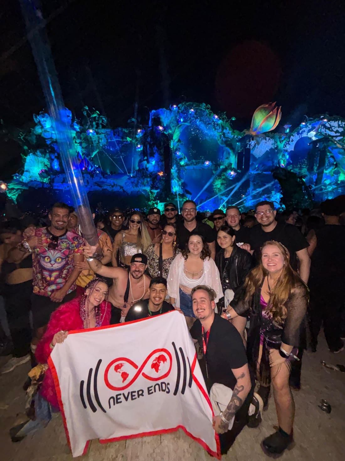 Grupo da Never Ends com a bandeira da agência diante do Mainstage da Tomorrowland Brasil iluminado de azul, à noite